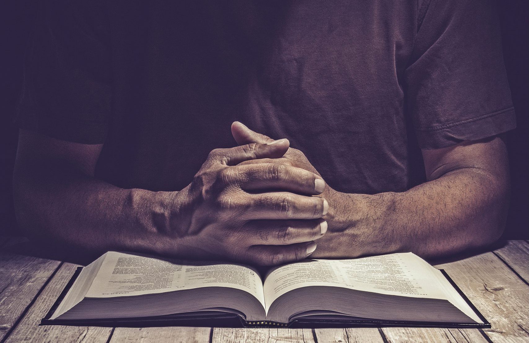 Man praying on a wooden table with an open Bible.